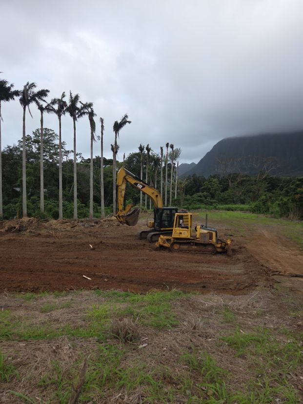 Excavator and dozer on a cleared dirt lot below an Oʻahu mountain ridge